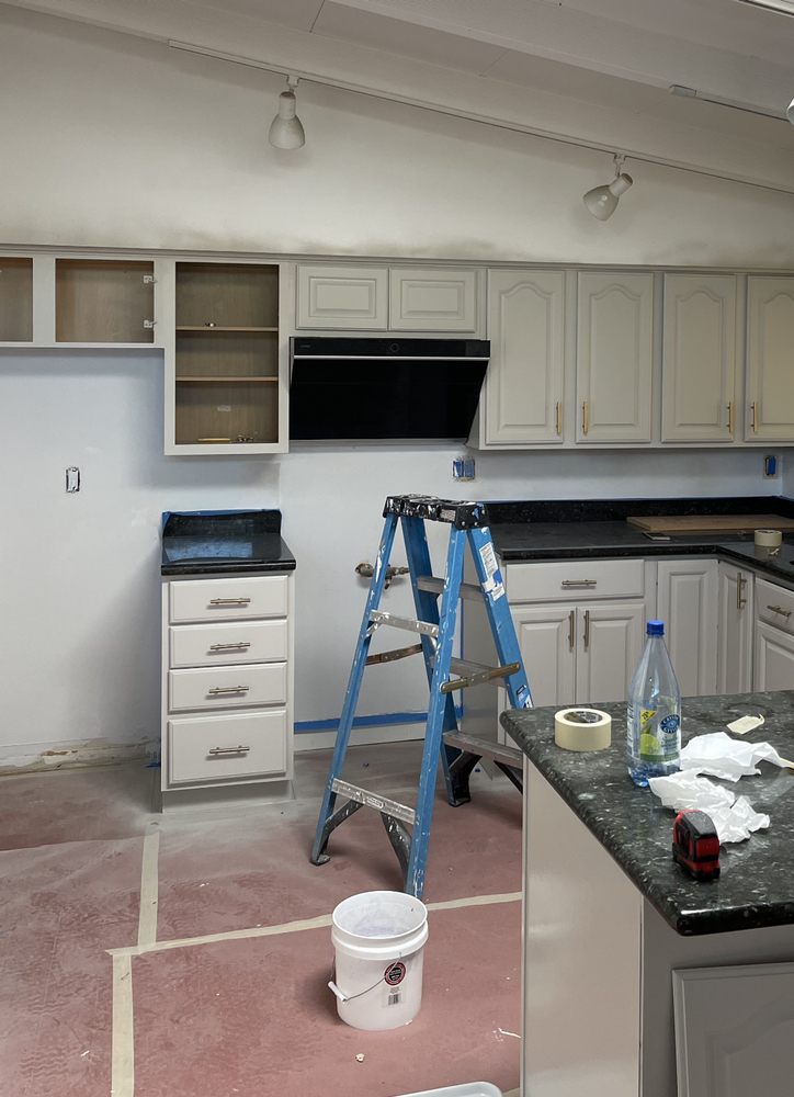 Modern kitchen under renovation with white cabinets, black countertops, and open shelves. A blue ladder and repainting tools are present, suggesting ongoing work.