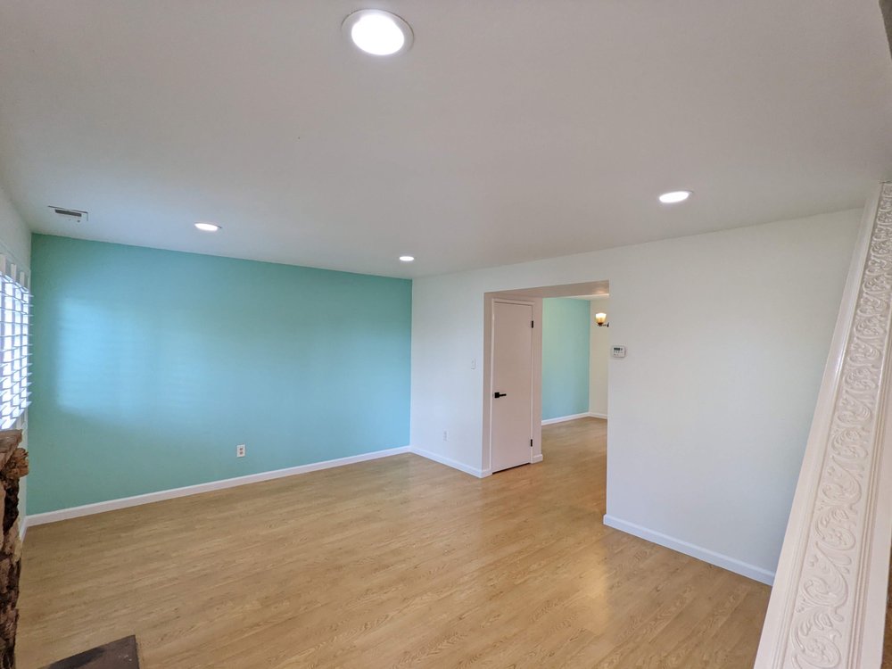 Spacious room with light wood flooring and white walls, featuring a turquoise accent wall. Ceiling lights create a bright and welcoming atmosphere.