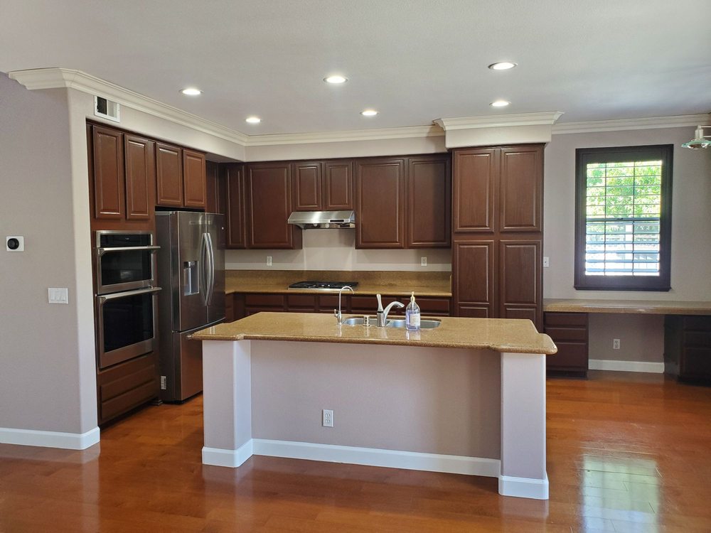 Spacious kitchen with dark wood cabinets, stainless steel appliances, and a large island with a sink. Warm lighting and a bright window create a cozy feel.